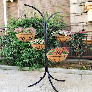 May include: A black metal hanging plant stand with four hanging baskets filled with colorful flowers. The baskets are made of a natural fiber material and are decorated with a decorative design. The stand is outdoors, in front of a green bush.