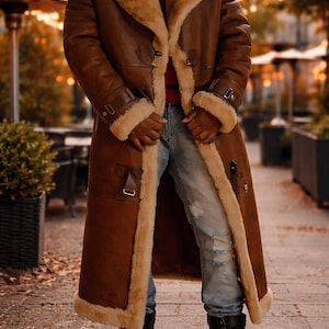 May include: A long, brown shearling coat with a tan shearling lining and trim. The coat features a wide collar, buckle details on the sleeves, and a knee-length design. The person is wearing blue jeans and black boots.