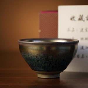 May include: A ceramic tea bowl with a dark, iridescent glaze, transitioning from gold to deep blue. A white card with Japanese text is behind the bowl, suggesting a certificate of authenticity. The bowl sits on a wooden surface.