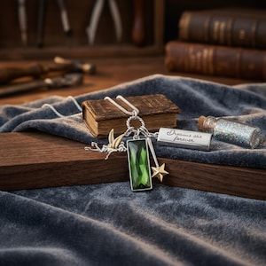 Puede incluir: Un collar de plata con un colgante rectangular de piedra verde, un charm de estrella dorada y una pequeña barra de plata grabada con "Dreams are forever". También hay una pequeña botella de vidrio llena de purpurina.