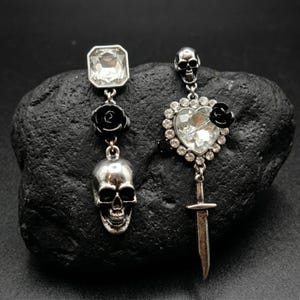 May include: A pair of mismatched earrings with a gothic theme. One earring features a silver skull pendant, a black rose, and a square crystal. The other earring has a heart-shaped crystal, a black rose, and a dagger pendant.