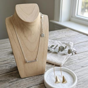 May include: A silver necklace with a bar pendant set with small, clear stones is displayed on a wooden jewelry bust. Matching earrings with gold triangle pendants are in a white bowl. The jewelry is on a light wood surface.