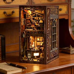 Puede incluir: Diorama de madera detallado de una cafetería y librería, iluminado desde el interior. La escena en miniatura incluye estanterías, mesas y pequeñas figuras. El exterior presenta el texto "SERENITY COFFEE HOUSE" y una ventana decorativa.