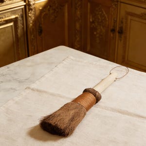 Op de afbeelding: Een houten gebakskwast met een bruine borstelkop en een gewikkelde handgreep. De kwast ligt op een beige linnen doek. De handgreep heeft een metalen lus om op te hangen.