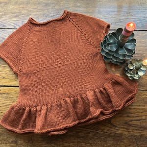 May include: A rust-colored, short-sleeved knitted top with a ruffled hem. The top is made of a soft, textured yarn and features a round neckline. The garment is displayed on a wooden surface, with decorative candle holders in the background.