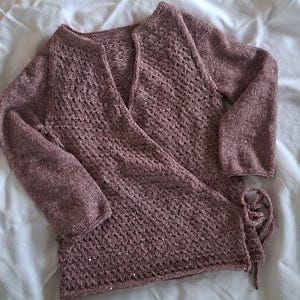 May include: A dusty rose wrap-style cardigan featuring long sleeves and a textured, open-knit pattern throughout the body. The garment includes a side tie closure with matching knitted strings and a V-neckline, crafted from a soft yarn with subtle shimmer.