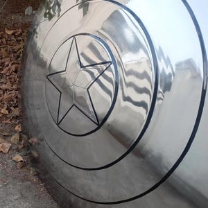 May include: A shiny, metallic shield with a prominent five-pointed star at its center. The shield has concentric circular bands and a black outline, creating a striking visual effect. The surface reflects the surrounding environment.