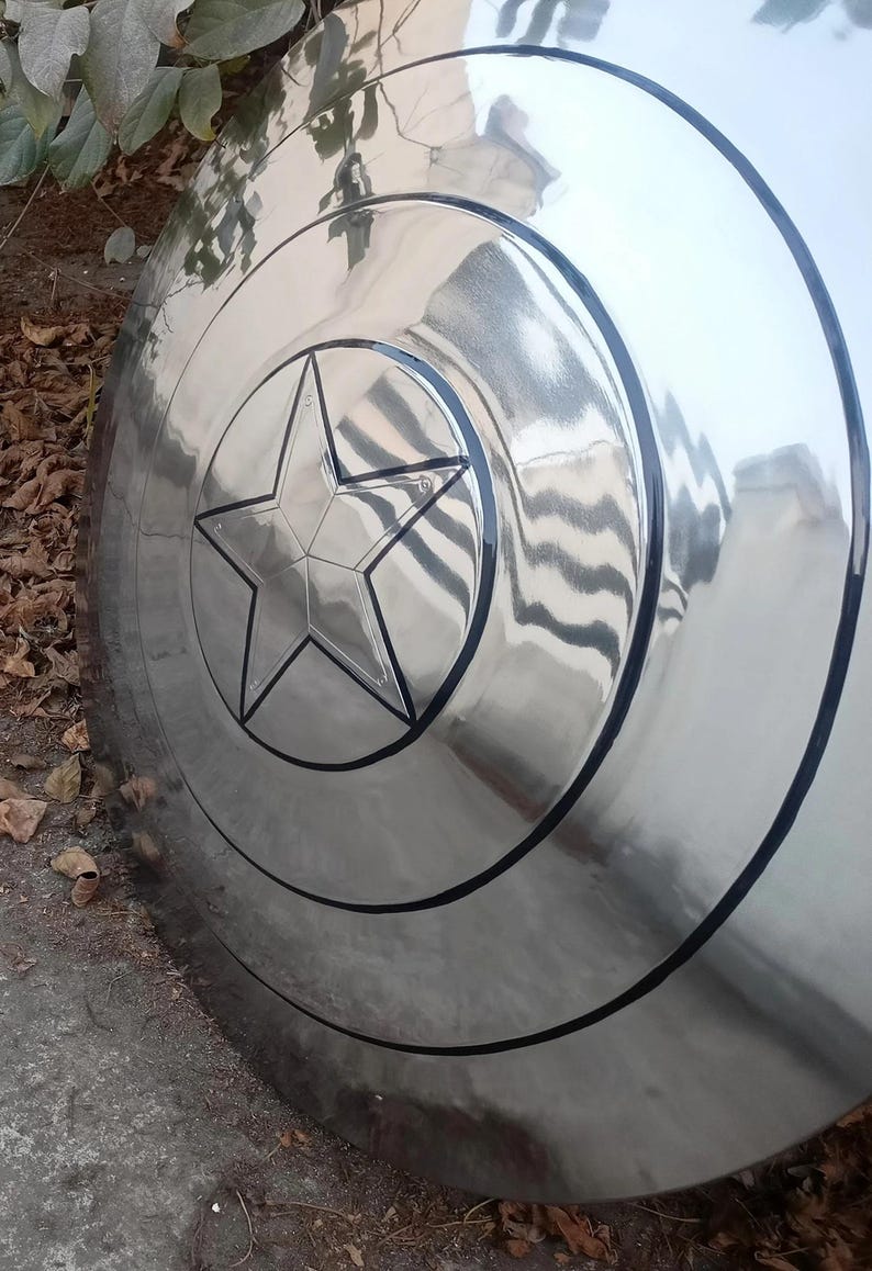 May include: A shiny, metallic shield with a prominent five-pointed star at its center. The shield has concentric circular bands and a black outline, creating a striking visual effect. The surface reflects the surrounding environment.