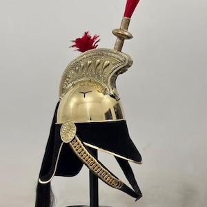Puede incluir: Un casco militar de color latón con visera de terciopelo negro y penacho rojo. El casco tiene intrincados detalles decorativos y una larga cola negra. El casco se exhibe sobre un soporte negro.