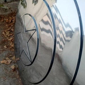 May include: A silver-colored shield with a prominent star emblem in the center. The shield has a circular design with a black border, reflecting light and appearing metallic. The star is also outlined in black.