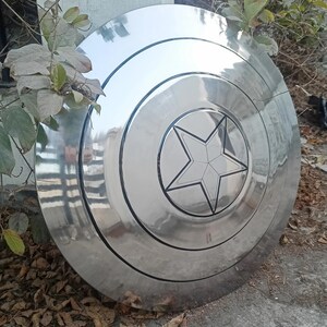 May include: A large, shiny, silver shield with a five-pointed star design in the center. The shield has concentric circular bands and a reflective surface. The shield is a prop or collectible.