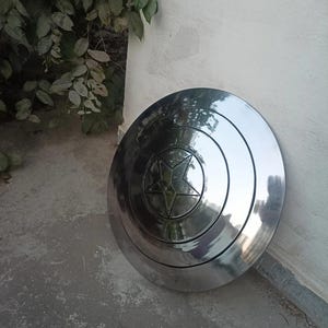 May include: A shiny, silver shield with a central star design. The shield is round with concentric circles and a five-pointed star in the center. The shield is leaning against a white wall.