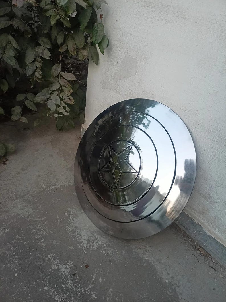 May include: A shiny, silver shield with a central star design. The shield is round with concentric circles and a five-pointed star in the center. The shield is leaning against a white wall.