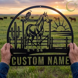 May include: Black metal wall art featuring a horse, an eagle, and a cactus within a circular frame. The design includes a fence, a rising sun, and the text "CUSTOM NAME" at the bottom. The background shows a green field and a sunset.