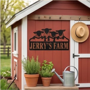 May include: A black metal sign featuring four sheep silhouettes above the text JERRY'S FARM in a bold font. The sign is mounted on the exterior wall of a red building, positioned above two terracotta pots with green plants and a metal watering can.