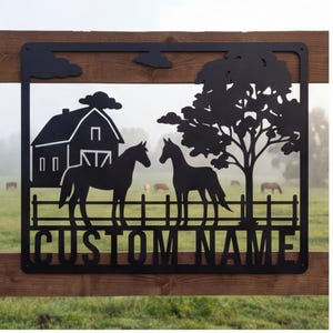 May include: A black metal wall sign featuring a cutout design of a barn, two horses, a tree, and a fence. The sign includes a rectangular frame and the text "CUSTOM NAME" in bold, capital letters at the bottom, showcasing a rural farm scene.
