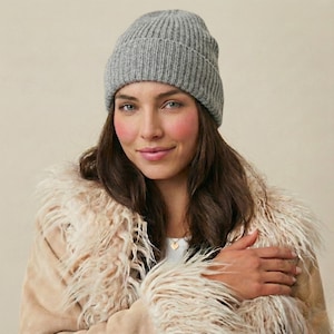 Könnte beinhalten: Eine graue, gerippte Strickmütze. Der Hut ist aus einem weichen, warmen Material gefertigt. Die Person trägt einen beigefarbenen Mantel mit einem flauschigen, cremefarbenen Besatz.
