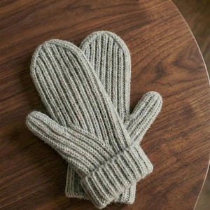 Può includere: Un paio di guanti in maglia grigio chiaro con un design a coste. I guanti sono realizzati in un materiale morbido e strutturato. Sono progettati per tenere le mani al caldo. I guanti sono su una superficie di legno.