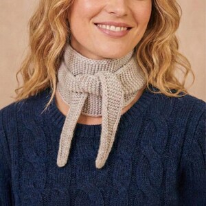 May include: A light grey knitted scarf tied in a knot, with long ends. The scarf is made of a thick, textured yarn. The person in the image is wearing a dark blue jumper.