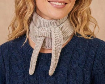 May include: A light gray knitted scarf tied in a knot, with long ends hanging down. The scarf is made of a thick, textured yarn. The person in the image is wearing a dark blue sweater.