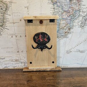 May include: A wooden wall-mounted key holder with a decorative metal emblem featuring a red tractor design and the letters "U" and "F". The key holder has two hooks and is mounted on a world map background.