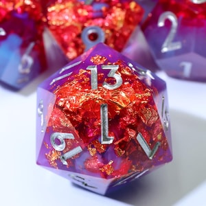 May include: A twenty-sided die made of translucent purple resin with embedded red and gold metallic foil flakes. The die features engraved numbers painted in silver, with the number 13 prominently displayed on the top triangular face.