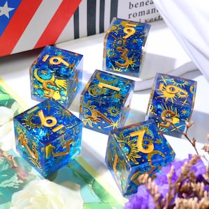 Könnte beinhalten: Ein Set aus fünf halbtransparenten blauen Harzwürfeln mit goldenen Himmelsmotiven wie Sonnen, Monden und Sternen. Jeder Würfel zeigt große, geprägte goldene Ziffern auf seinen Seiten und ein glitzerndes, tiefblaues Inneres durch das klare Material.
