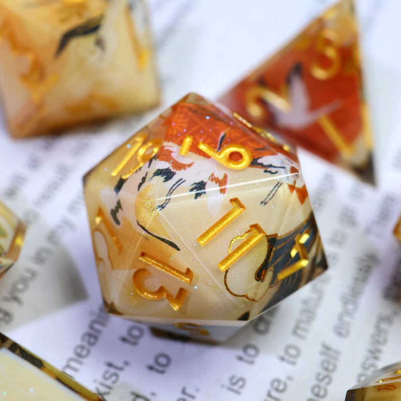 May include: Close-up of a set of polyhedral dice. The dice are made of clear resin with a cream-coloured fabric insert featuring a floral and bird design. The numbers are gold. The dice are for tabletop games.