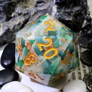 May include: A large, multi-sided die with a floral and wave pattern in shades of blue, green, and pink. The numbers are gold. The die is set against a background of rocks and white and black stones.