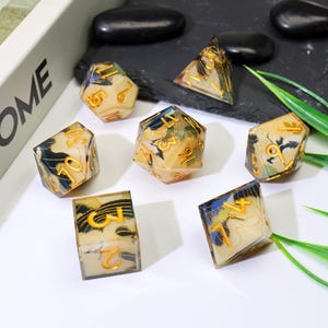 May include: A collection of seven polyhedral dice. Each die has a cream-coloured base, gold numbering, and a decorative pattern with blue and black accents. The dice are displayed on a dark surface with green foliage.