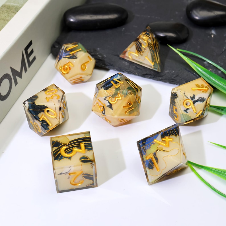 May include: A collection of seven polyhedral dice. Each die has a cream-coloured base, gold numbering, and a decorative pattern with blue and black accents. The dice are displayed on a dark surface with green foliage.