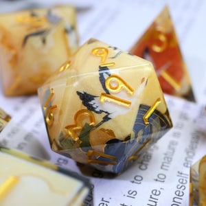 May include: A set of polyhedral dice with a Japanese-inspired design. The dice are clear with a cream-coloured interior featuring a crane and floral pattern. Gold numbers and accents are visible. Other dice are in the background.