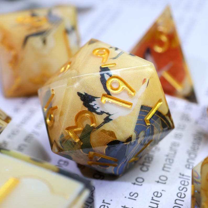 May include: A set of polyhedral dice with a Japanese-inspired design. The dice are clear with a cream-coloured interior featuring a crane and floral pattern. Gold numbers and accents are visible. Other dice are in the background.