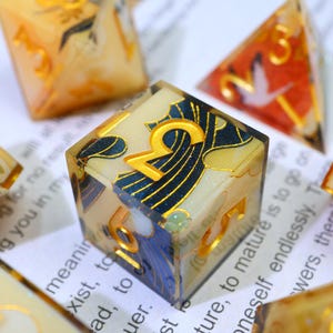 May include: Close-up of a set of decorative dice. The dice are clear with gold numbers and intricate designs in blue, gold, and white. The dice are on a white surface with printed text.