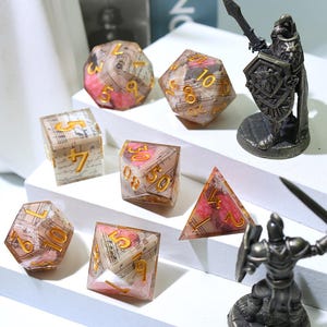 May include: A collection of polyhedral dice with a musical theme. The dice are clear with pink and white accents, and gold-colored numbers. Two small metal figurines of knights are also present.