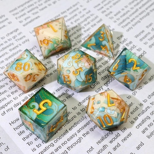 May include: A collection of seven polyhedral dice with a floral and wave design. The dice feature a mix of colors, including teal, gold, and cream. Each die has gold numbers. The dice are designed for tabletop games.