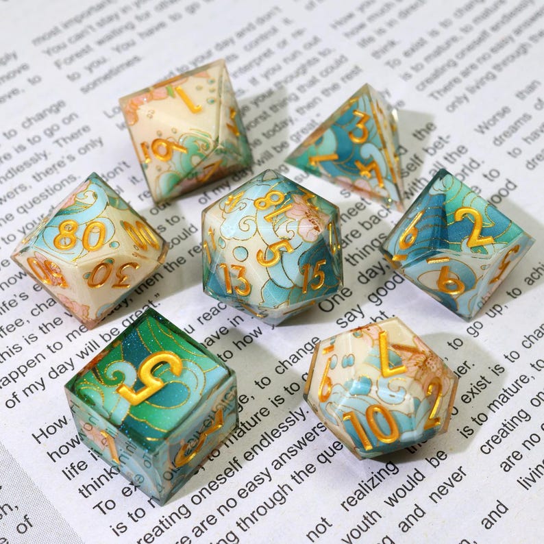 May include: A collection of seven polyhedral dice with a floral and wave design. The dice feature a mix of colors, including teal, gold, and cream. Each die has gold numbers. The dice are designed for tabletop games.