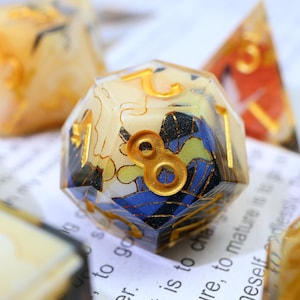 May include: A clear resin die with multiple sides, displaying gold numerals and an intricate interior design with blue, yellow, and black patterns. The number 8 is highlighted in gold. Other dice are visible in the background.