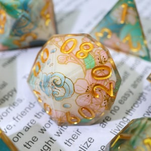 May include: A multi-sided die with a floral design. The die features a cream-colored base with pink and blue floral patterns and gold numbering. Other dice with similar designs are visible in the background.