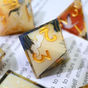May include: Close-up of a set of polyhedral dice. The dice are clear with gold numbers and decorative designs. The edges are outlined in dark blue. The dice are resting on a white surface with printed text.
