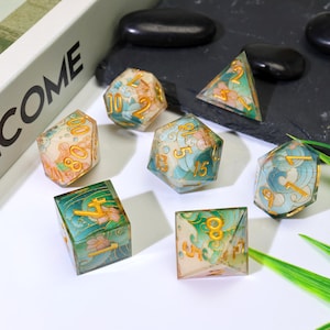 May include: A set of seven polyhedral dice with a floral and wave design. The dice are clear with gold numbering and accents, featuring shades of green, blue, and peach. The dice are arranged on a dark surface with a white background.