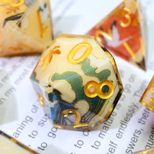 May include: A set of polyhedral dice with a cream-coloured base and gold numbering. The dice feature a Japanese-inspired design with green and blue accents. The numbers are in a gold colour. The dice are on a white surface with text.