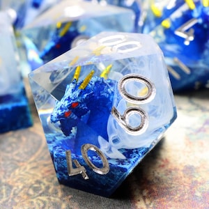 May include: A translucent multi-sided die featuring a blue dragon figurine with yellow horns and red eyes encased inside. The die has white swirling smoke-like patterns and silver-colored numbers including 40, 60, and 00 engraved on the exterior faces.