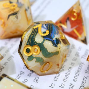 May include: A set of decorative polyhedral dice. The dice have a cream-coloured base, gold numbering, and intricate designs in green, blue, and gold. The dice have a Japanese-inspired aesthetic, with detailed artwork.