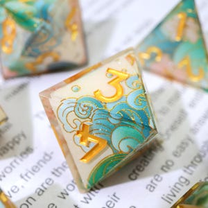 May include: Close-up of a set of polyhedral dice. The dice are clear with a cream-colored interior and gold numbers. Each die features a blue and green wave design with gold accents. The number "3" is prominently displayed on one die.