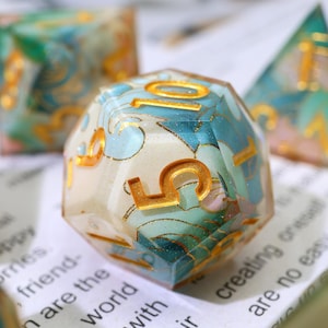 May include: A multi-sided, clear resin die with gold numbering. The die is filled with a blue and white floral pattern. Other dice are visible in the background. The number 5 is prominently displayed on the front of the die.