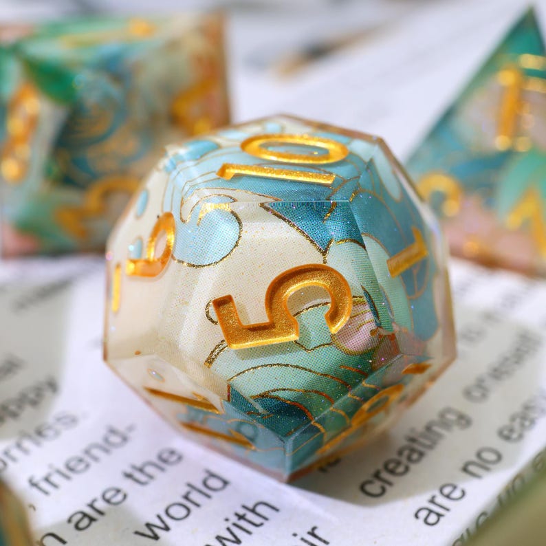 May include: A multi-sided, clear resin die with gold numbering. The die is filled with a blue and white floral pattern. Other dice are visible in the background. The number 5 is prominently displayed on the front of the die.