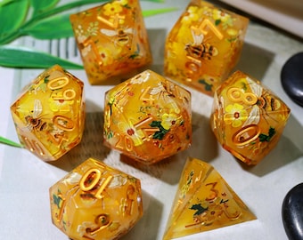May include: Set of seven polyhedral dice with a yellow and orange resin base. Each die features embedded bees, flowers, and gold numbering. The dice are designed for tabletop games and have a unique, artistic appearance.