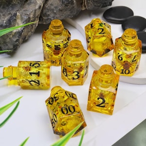 May include: A collection of yellow resin dice with black numbers and insect inclusions. The dice are in various shapes, including rectangular and hexagonal, and are designed for tabletop gaming.