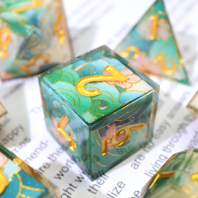 May include: Close-up of a set of dice with a floral and wave design. The dice are a mix of green, blue, and pink with gold-colored numbers. The dice are on a white surface with printed text.